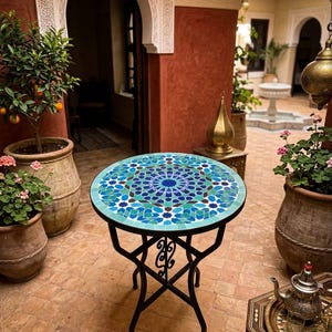 Könnte beinhalten: Ein runder Mosaiktisch mit schwarzem Metallgestell. Die Tischplatte weist ein geometrisches Muster in Blau, Türkis und Braun auf. Der Tisch befindet sich im Freien, umgeben von Topfpflanzen und architektonischen Details.