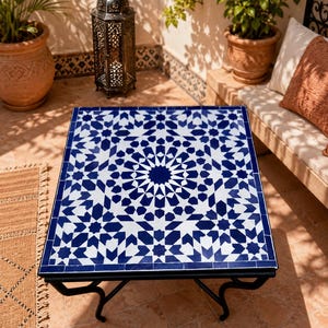 May include: A square table featuring a white and blue geometric tile mosaic top with a star-patterned design. The table is supported by a black metal frame with four curved legs that extend outward from the center to provide a stable base.