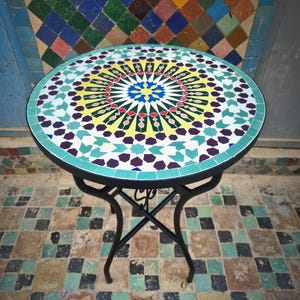 May include: A round mosaic tile table with a black metal base. The tabletop features a colorful geometric pattern in shades of blue, yellow, green, and purple. The table is approximately 24 inches in diameter.