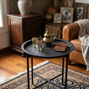 May include: A black metal side table with a decorative tray top holds a gold teapot, two glasses, and a leather-bound book. The table sits on a patterned rug. The table's height is approximately 60 cm.