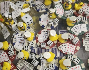 Puede incluir: Una colección de artículos novedosos con patos de goma amarillos, dados, naipes y billetes en miniatura. Algunos patos llevan sombreros. Los artículos están dispuestos sobre bases en forma de fichas de póquer. También se ven llaveros con etiquetas.
