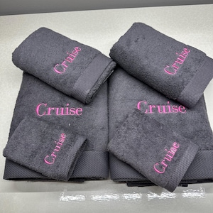 May include: A set of dark gray towels, including bath towels and washcloths, embroidered with the name "Cruise" in pink. The towels are neatly folded and stacked, showcasing a coordinated bathroom set.