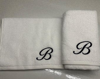 May include: Two white cotton towels, one folded and one rolled, with a black embroidered letter "B". The towels are displayed on a light gray surface. The towels are a bathroom or spa item.
