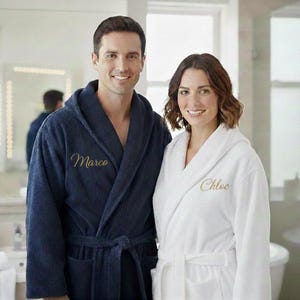 May include: A couple stands in a bathroom, each wearing a personalized bathrobe. The man wears a navy blue robe embroidered with "Marco" in gold script. The woman wears a white robe embroidered with "Chloe" in gold script. Both robes have hoods and waist ties.