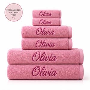 May include: A stack of six pink terry cloth towels of varying sizes, each embroidered with the name Olivia in a dark pink cursive script. The towels are arranged in a pyramid shape, with the smallest at the top and the largest at the bottom.
