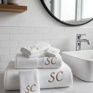May include: A stack of white bath towels with gold embroidered "SC" initials. The towels are arranged on a white countertop in a bathroom setting. The bathroom has a white sink, a mirror, and a small plant on a shelf.