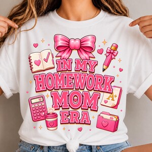 Może przedstawiać: Biały t-shirt z różowym nadrukiem graficznym i napisem "IN MY HOMEWORK MOM ERA". Wzór zawiera dużą różową kokardę, notatnik, ołówek, kalkulator, kubek kawy, teczkę i kartkę z oceną A+, wszystko ozdobione małymi serduszkami.