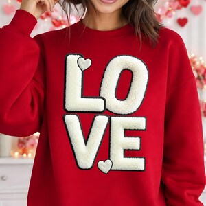 May include: A bright red crewneck sweatshirt featuring the word LOVE in large, cream-coloured, fuzzy, sherpa-style letters. The letters are outlined in black, and two small, matching cream-coloured fuzzy hearts are positioned near the letters.