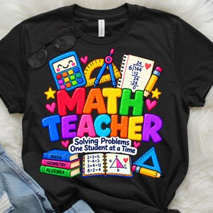 Può includere: Una t-shirt nera con una grafica colorata e il testo "MATH TEACHER Solving Problems One Student at a Time". Il design include una calcolatrice, un goniometro, equazioni matematiche, libri etichettati "MATH", "GEOMETRY", "ALGEBRA" e un righello.