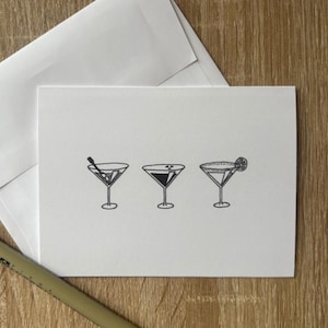 Può includere: Un biglietto di auguri bianco con tre bicchieri da martini illustrati con inchiostro nero. Ogni bicchiere ha una guarnizione diversa: un'oliva, un liquido scuro e una fetta di limone. Una busta e una penna sono sullo sfondo.