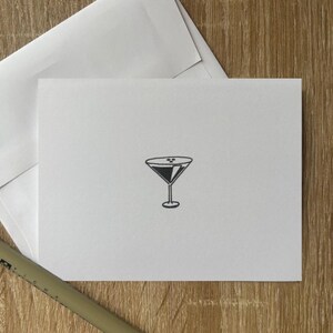 Può includere: Un biglietto di auguri bianco con un'illustrazione di un bicchiere da martini nero. Il biglietto è accompagnato da una busta bianca e una penna nera, il tutto su una superficie di legno. Il bicchiere da martini ha un gambo e un bicchiere triangolare.