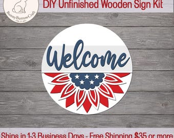 May include: A circular wooden sign kit featuring the word Welcome in dark blue script. Below the text is a stylized sunflower graphic with red petals and a dark blue center adorned with white stars, reflecting a patriotic color scheme.