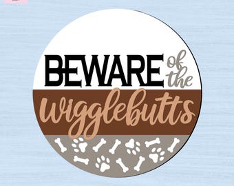 May include: A circular sign featuring a white top section with the black text BEWARE of the, a brown middle section with the light brown text wigglebutts, and a gray bottom section decorated with white paw prints and bone shapes throughout.
