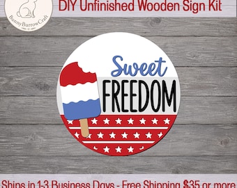 May include: A circular wooden sign featuring a red, white, and blue popsicle graphic on the left. The text "Sweet FREEDOM" is printed in blue and black. The lower portion of the sign displays a red background with white stars and horizontal lines.