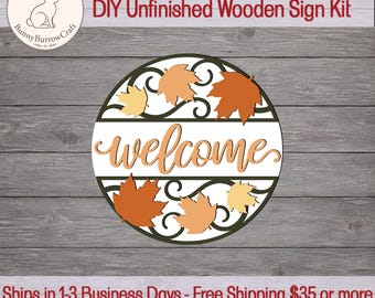 May include: A circular DIY unfinished wooden sign kit featuring a cutout design with dark green scrollwork and light orange, orange, and dark orange maple leaves. The word welcome is written in a cursive script across the center of the sign.