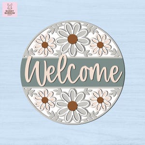 May include: Round wooden welcome sign with a daisy flower design. The sign features the word "Welcome" in a cursive font, set against a sage green background. The daisies have gray petals and brown centers. The sign is a decorative item.