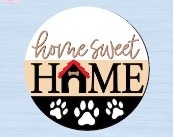 May include: A circular wooden sign featuring the text "home sweet HOME" in black and brown script and block lettering. The letter "O" in the word "HOME" is replaced by a black dog house with a red roof and a small bone icon above the entrance.