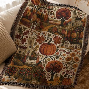 May include: A woven throw blanket featuring a detailed autumn landscape with pumpkins, colorful fall foliage, trees, and traditional houses. The design includes a large central pumpkin, apple trees, and a church tower, finished with a fringed edge.