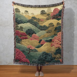 May include: A woven throw blanket featuring a landscape design with rolling hills, trees, and a small building. The color palette includes shades of green, gold, and red. The blanket has a fringed edge.