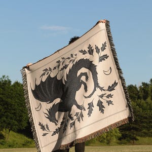 May include: A beige throw blanket with a black dragon design, oak leaves, and crescent moons. The blanket has a fringed edge and is held up against a blue sky and green trees.