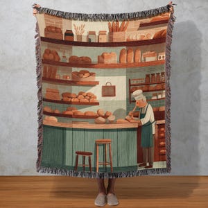 May include: A woven throw blanket featuring a bakery scene. The illustration depicts a baker in a white hat and apron, surrounded by shelves of bread and baked goods. The color palette includes shades of brown, green, and beige.