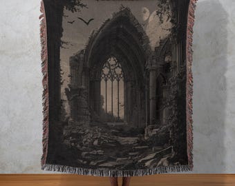 May include: A black and grey throw blanket depicting a gothic cathedral scene with bats, a full moon, and a detailed arched window. The blanket has a fringed edge and is held up against a neutral background.