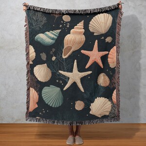 May include: A dark blue throw blanket featuring a pattern of seashells and starfish in shades of beige, peach, and teal. The blanket has a fringed edge and is held up by a person. The design includes various shell types and coral.