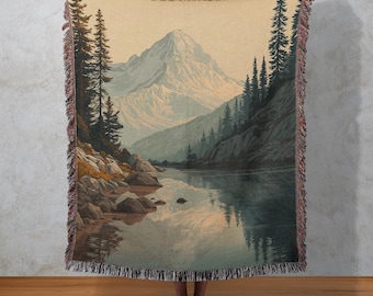 Può includere: Una coperta tessuta con un paesaggio panoramico con una montagna, un lago e alberi. La coperta ha uno sfondo color crema con un'illustrazione dettagliata in tonalità di verde, blu e marrone. I bordi sono rifiniti con una frangia.