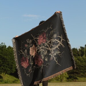 May include: A black throw blanket with a floral design of roses and thorns. The blanket has a fringed edge and is held up against a blue sky. The roses are in shades of red and pink, with a brown fringe.