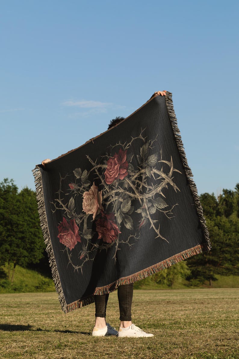 May include: A black throw blanket with a floral design of roses and thorns. The blanket has a fringed edge and is held up against a blue sky. The roses are in shades of red and pink, with a brown fringe.