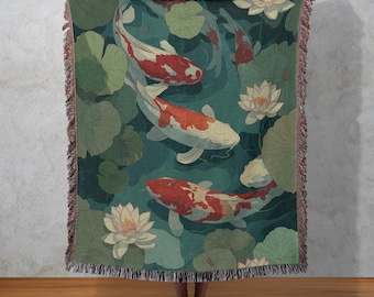 May include: A woven throw blanket featuring a koi pond design. The blanket displays koi fish with red and white markings swimming among lily pads and lotus flowers in shades of green and teal. The blanket has a fringed edge.