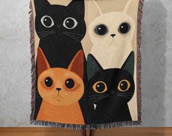 May include: A woven throw blanket featuring a whimsical design of four cartoon cats. The cats are black, orange, white, and black, each with large, expressive eyes. The blanket has a fringed edge and a beige background.