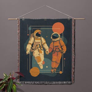 May include: A woven wall hanging featuring two astronauts holding hands in space. The astronauts are in cream and orange space suits against a dark blue background with orange and yellow planets. The wall hanging is displayed with a wooden rod and a clear plastic hanger.