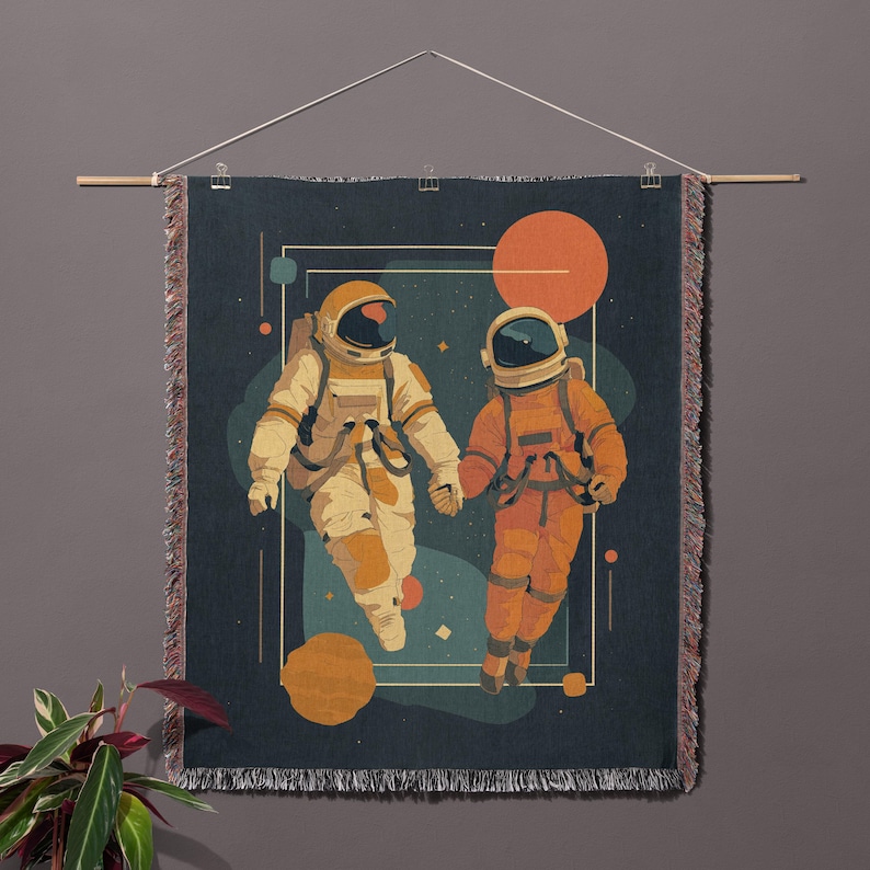 May include: A woven wall hanging featuring two astronauts holding hands in space. The astronauts are in cream and orange space suits against a dark blue background with orange and yellow planets. The wall hanging is displayed with a wooden rod and a clear plastic hanger.