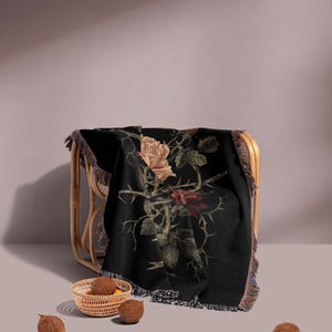 May include: A black throw blanket with a rose design in beige, red, and green, draped over a wicker chair. The blanket has a fringed edge. A small woven basket with brown objects and three coconuts are in the foreground.