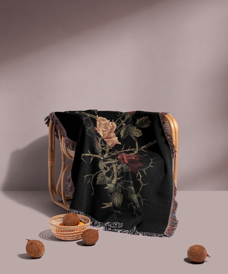 May include: A black throw blanket with a rose design in beige, red, and green, draped over a wicker chair. The blanket has a fringed edge. A small woven basket with brown objects and three coconuts are in the foreground.