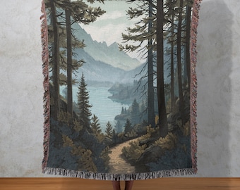 Può includere: Coperta tessuta raffigurante un paesaggio con un sentiero che conduce a un lago circondato da alberi e montagne. La tavolozza dei colori comprende sfumature di verde, blu e marrone, con un bordo a frange.