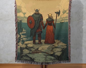 May include: Woven throw blanket featuring a Viking couple standing on a cliff overlooking the sea. The artwork depicts a man with a shield and a woman with an axe, holding hands. The color palette includes shades of orange, green, and blue.