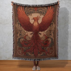 May include: A woven throw blanket featuring a detailed illustration of a phoenix with outstretched wings. The bird is primarily red with brown and tan accents, set against a backdrop of ornate scrollwork. The blanket has a fringed edge.