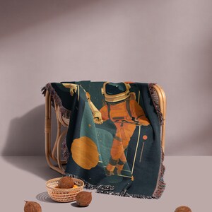 May include: A dark teal throw blanket with a fringed edge, featuring an astronaut in an orange spacesuit against a backdrop of stars and a large orange planet. The blanket is draped over a wicker chair.