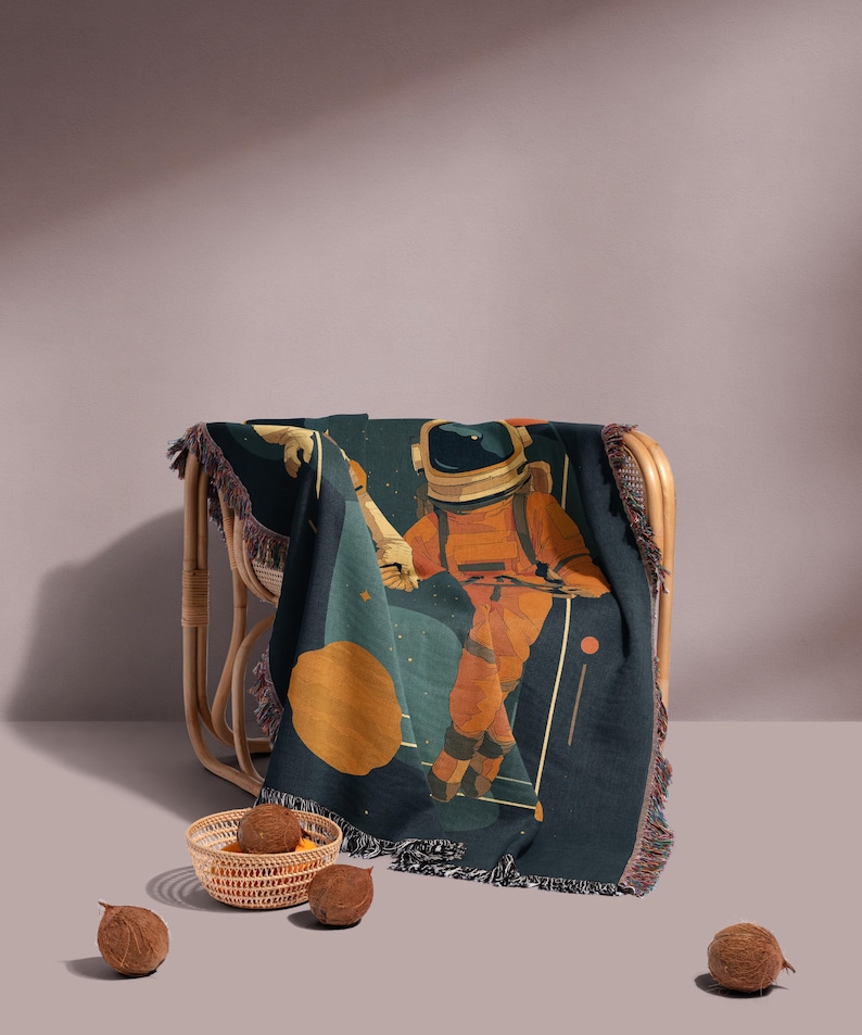 May include: A dark teal throw blanket with a fringed edge, featuring an astronaut in an orange spacesuit against a backdrop of stars and a large orange planet. The blanket is draped over a wicker chair.