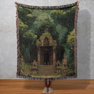 Könnte beinhalten: Eine gewebte Decke mit einer detaillierten Illustration eines alten Tempels, umgeben von üppigen grünen Bäumen. Das Kunstwerk zeigt steinerne Statuen und Stufen, die zu einem dunklen Tor führen, mit einem Fransenrand. Die Farbpalette umfasst Grün-, Braun- und Grautöne.