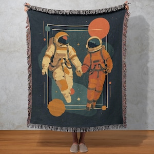 May include: A woven throw blanket featuring an illustration of two astronauts holding hands in space. The astronauts are wearing orange and cream-colored space suits. The background is dark blue with orange and teal planets.