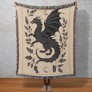 May include: Beige woven throw blanket featuring a black dragon design with outstretched wings, surrounded by leaves and crescent moons. The blanket has a fringed edge and is held up by two people.