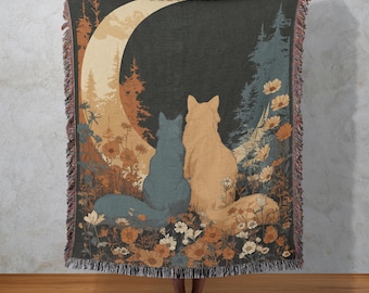 May include: A woven throw blanket featuring a whimsical illustration of two fox-like creatures gazing at a crescent moon. The design includes a forest scene with flowers in shades of blue, orange, and cream. The blanket has a fringed edge.