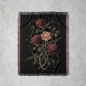 May include: Black throw blanket with a botanical illustration of roses and thorns. The roses are in shades of red and peach, with green leaves and brown thorns. The blanket has a fringed edge.