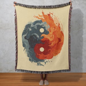 May include: Cream-coloured throw blanket with a Yin and Yang design. The design shows fire and water in shades of red, orange, and blue. The blanket has a fringed edge and is held up by a person.