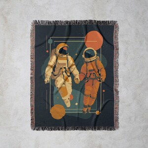 May include: A woven throw blanket featuring an illustration of two astronauts holding hands in space. The astronauts are in orange and beige space suits, set against a dark blue background with planets and stars. The blanket has a fringed edge.