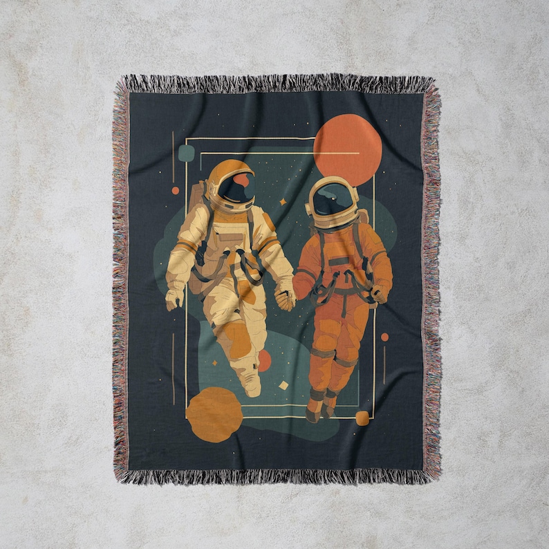 May include: A woven throw blanket featuring an illustration of two astronauts holding hands in space. The astronauts are in orange and beige space suits, set against a dark blue background with planets and stars. The blanket has a fringed edge.