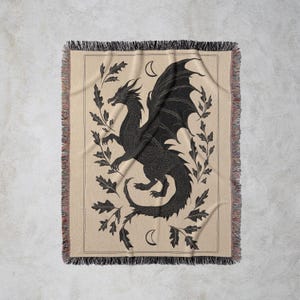 May include: A beige woven throw blanket featuring a black dragon illustration with outstretched wings, surrounded by oak leaves and crescent moons. The blanket has a fringed edge.